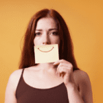 woman holding yellow sticky to face with red smile drawn on