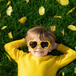 Child wearing heart shaped yellow glasses with yellow shirt, hands behind head smiling at camera laying on grass