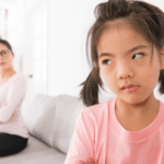 toddler girl looking frustrated as mother sits on couch behind her